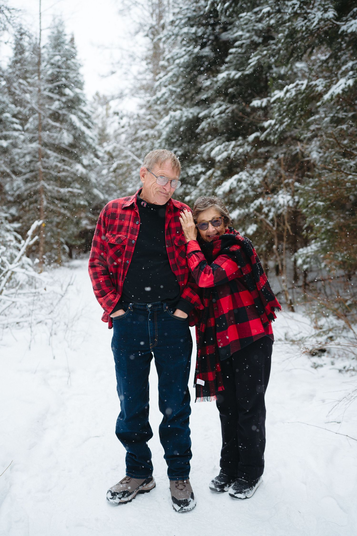 Two people wearing matching red buffalo plaid jackets pose together on a snowy winter trail surrounded by evergreen trees.