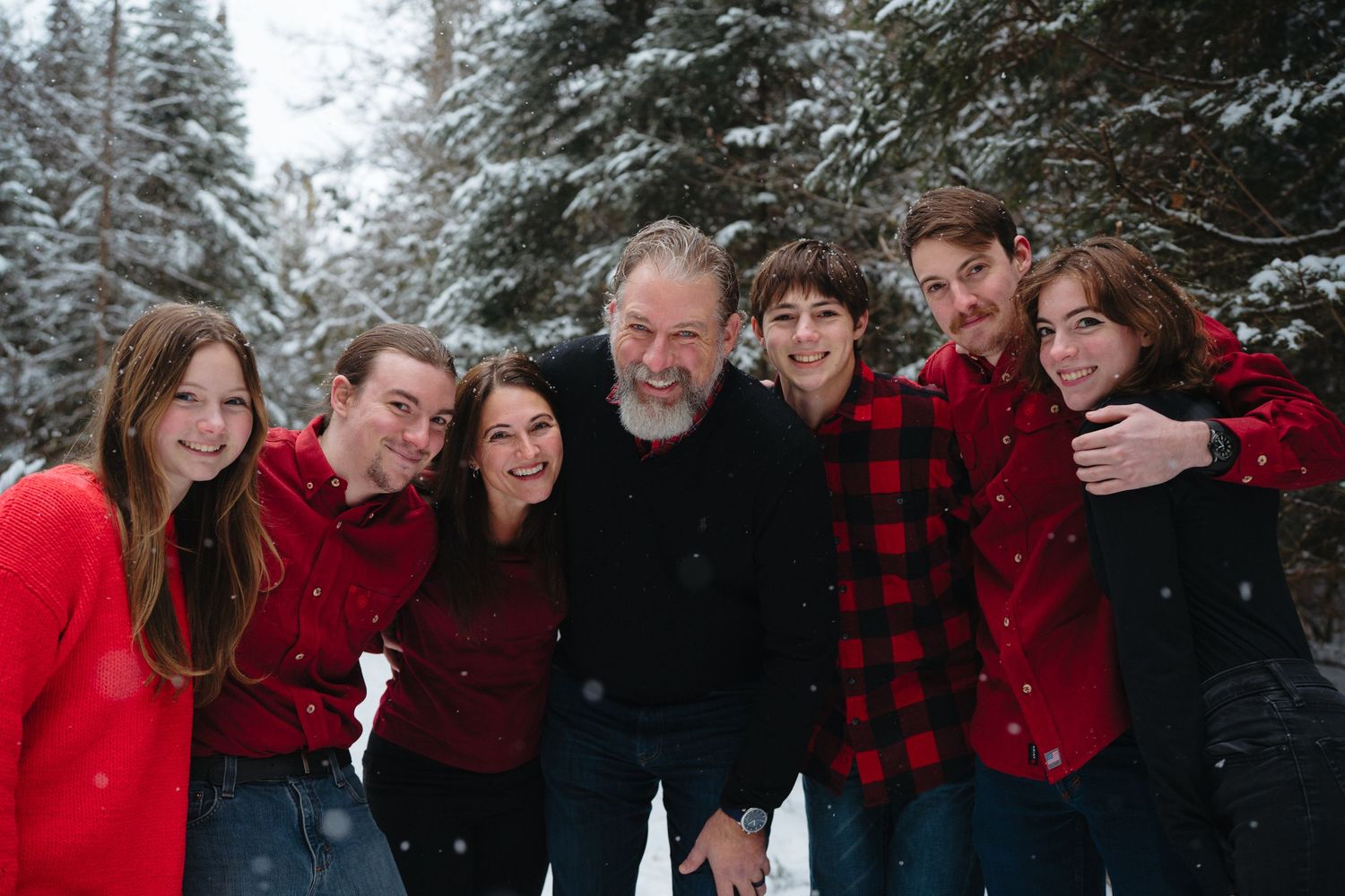 A group wearing coordinated red and black buffalo plaid outfits poses together in a snowy winter forest setting.