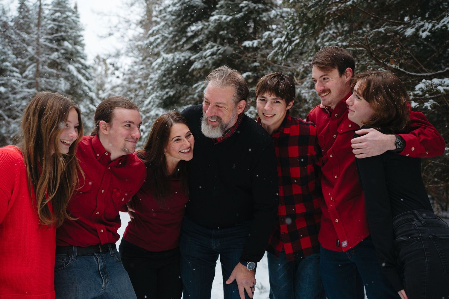 A family wearing coordinated red and black outfits poses together in a snowy forest setting during winter.