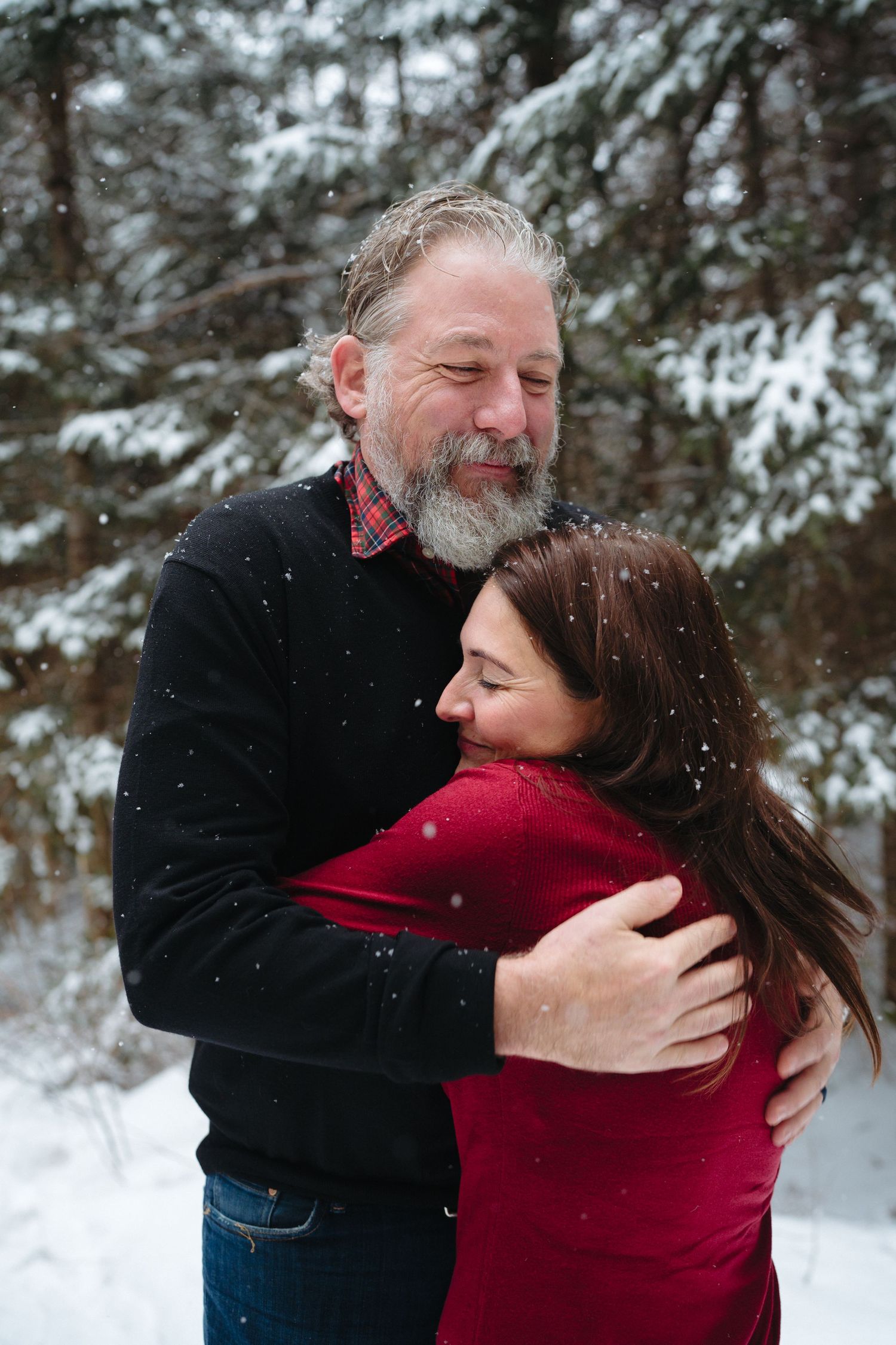 A couple embraces in a snowy winter forest setting wearing red and black clothing.