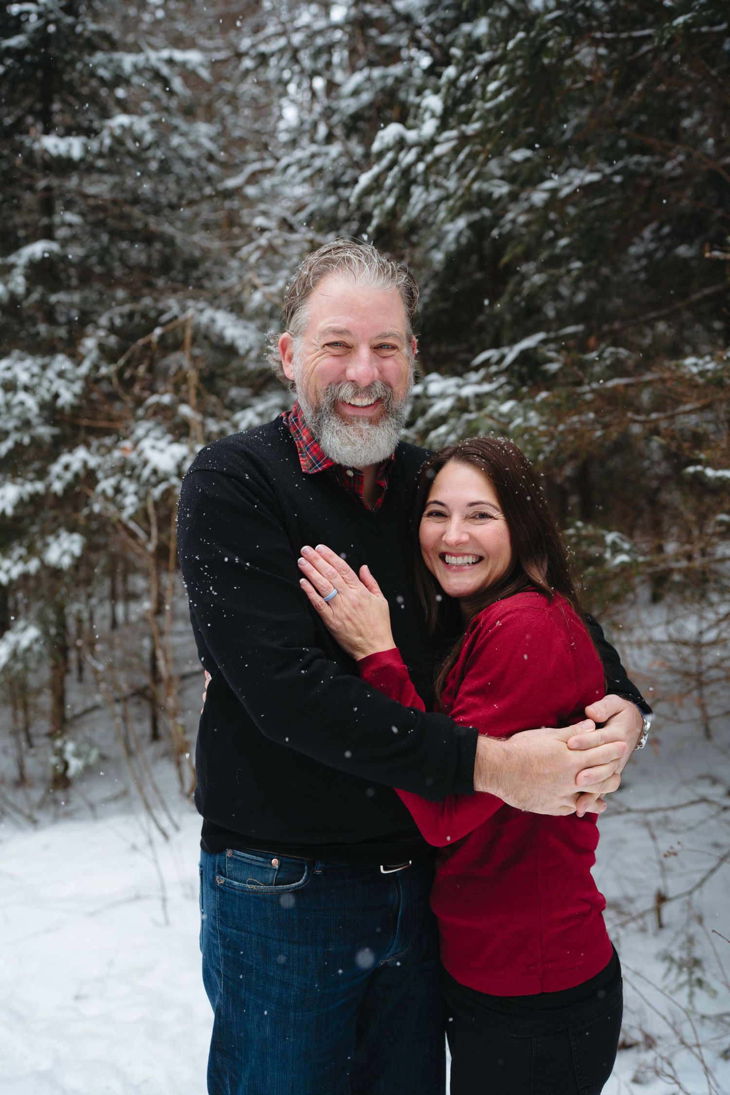 Two people share joyful laughter while hugging outdoors in a snow-covered forest landscape.