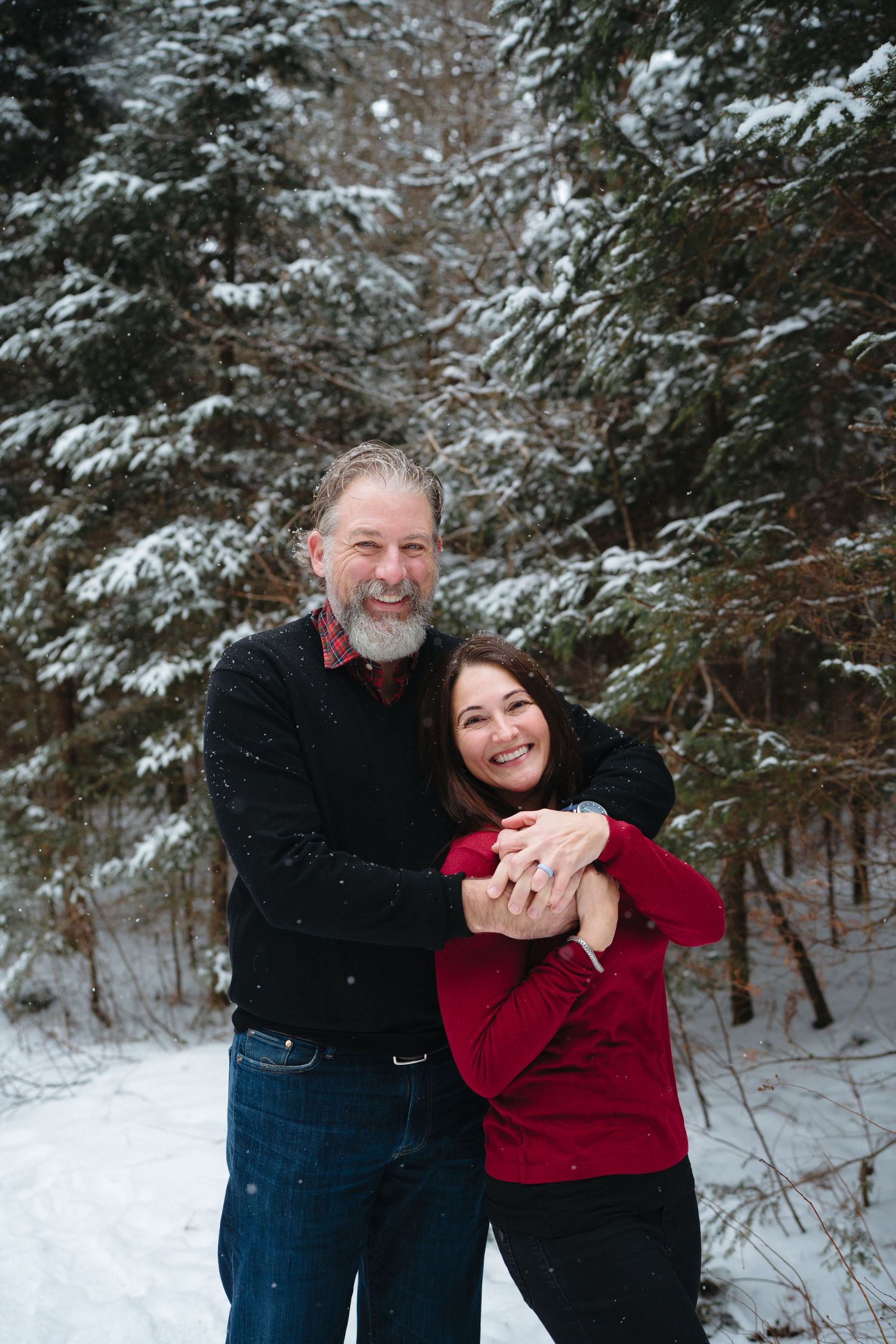 Two people share a playful embrace while wearing winter clothing against a snowy evergreen forest backdrop.