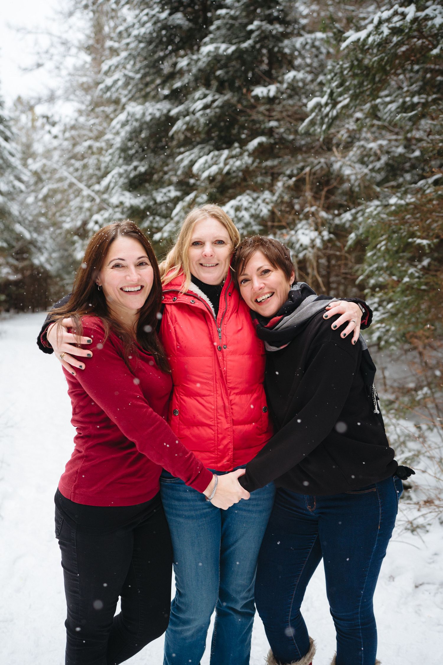 Three people wearing winter coats embrace and laugh together in a snowy forest setting.