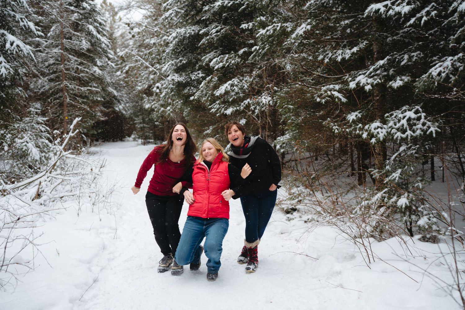 Three people walking together and laughing on a snow-covered forest path lined with evergreen trees.