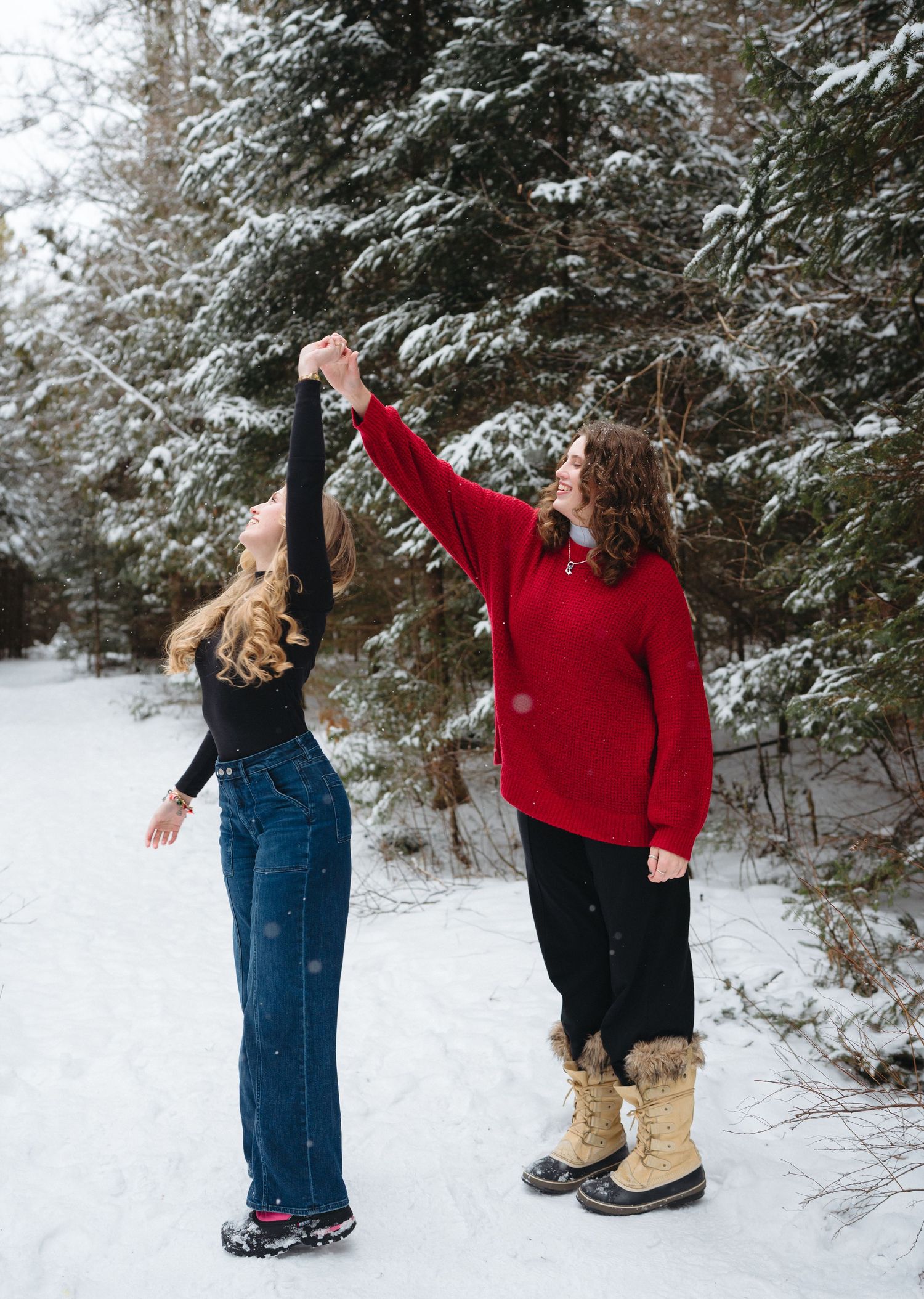 People dance and twirl playfully in the snow surrounded by evergreen trees.