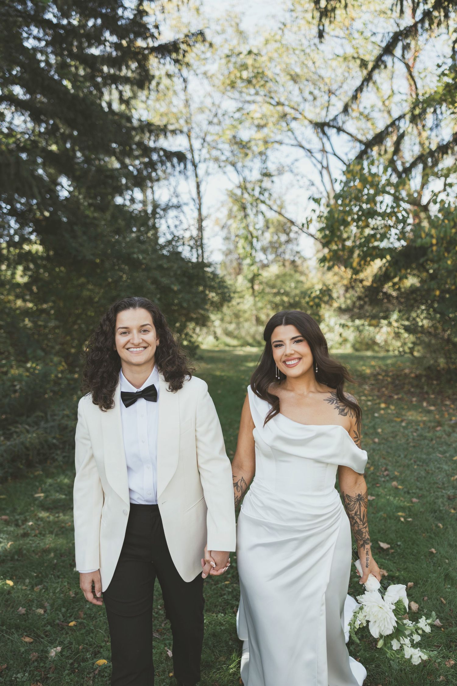 Two people in wedding attire smile and hold hands while standing in a sunny outdoor garden setting.