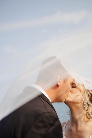 A romantic kiss under a flowing white veil against a soft blue sky background.