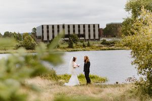 A couple stands together by a serene lake with a modern striped building in the background on a cloudy day.