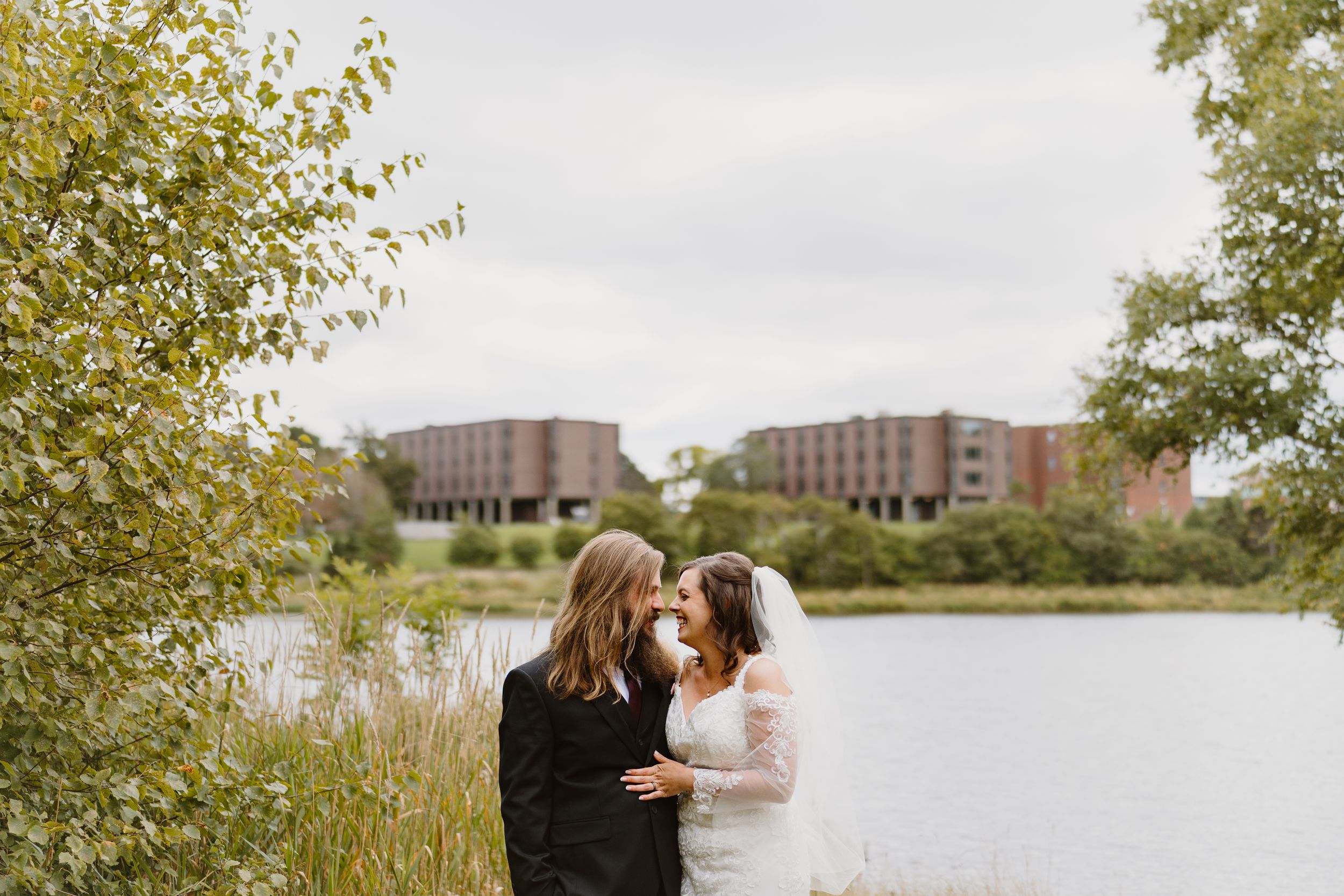A couple stands together in an autumn field near a lake with brick buildings visible in the background.
