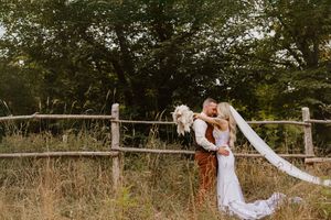 A romantic outdoor wedding photo at sunset with a couple embracing by a rustic wooden fence in a field.