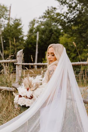 A bride wearing a flowing white veil and dress poses joyfully in a natural outdoor setting with tall grass and trees.