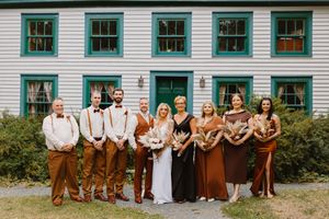 A wedding party poses in front of a white building wearing brown and rust colored outfits in a rustic outdoor setting.