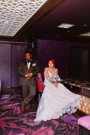 A bride in a lace gown and groom in a tuxedo pose elegantly in a purple-themed venue.