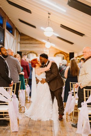 Newlywed interracial couple sharing first kiss at wedding ceremony aisle in rustic barn venue with guests standing.