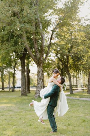 A romantic wedding couple embraces in a sweeping dance pose under a canopy of trees in a sunlit park.