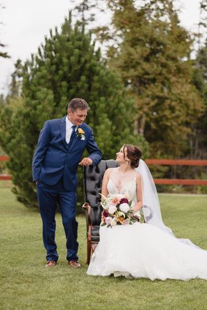 A couple in wedding attire sitting together on grass surrounded by evergreen trees.