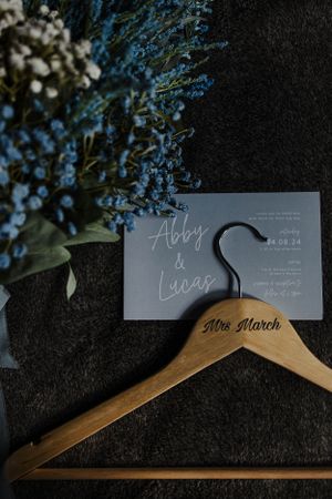 Wedding invitation and wooden hanger with 'Mrs March' text displayed with blue flowers against dark background.