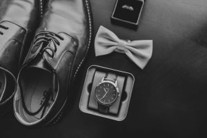 Close-up details of wedding accessories including shoes, rings, and tie arranged on dark surface.