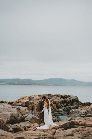 Bride and groom stand on rocky coastal shoreline in wedding attire with ocean and mountains in background.