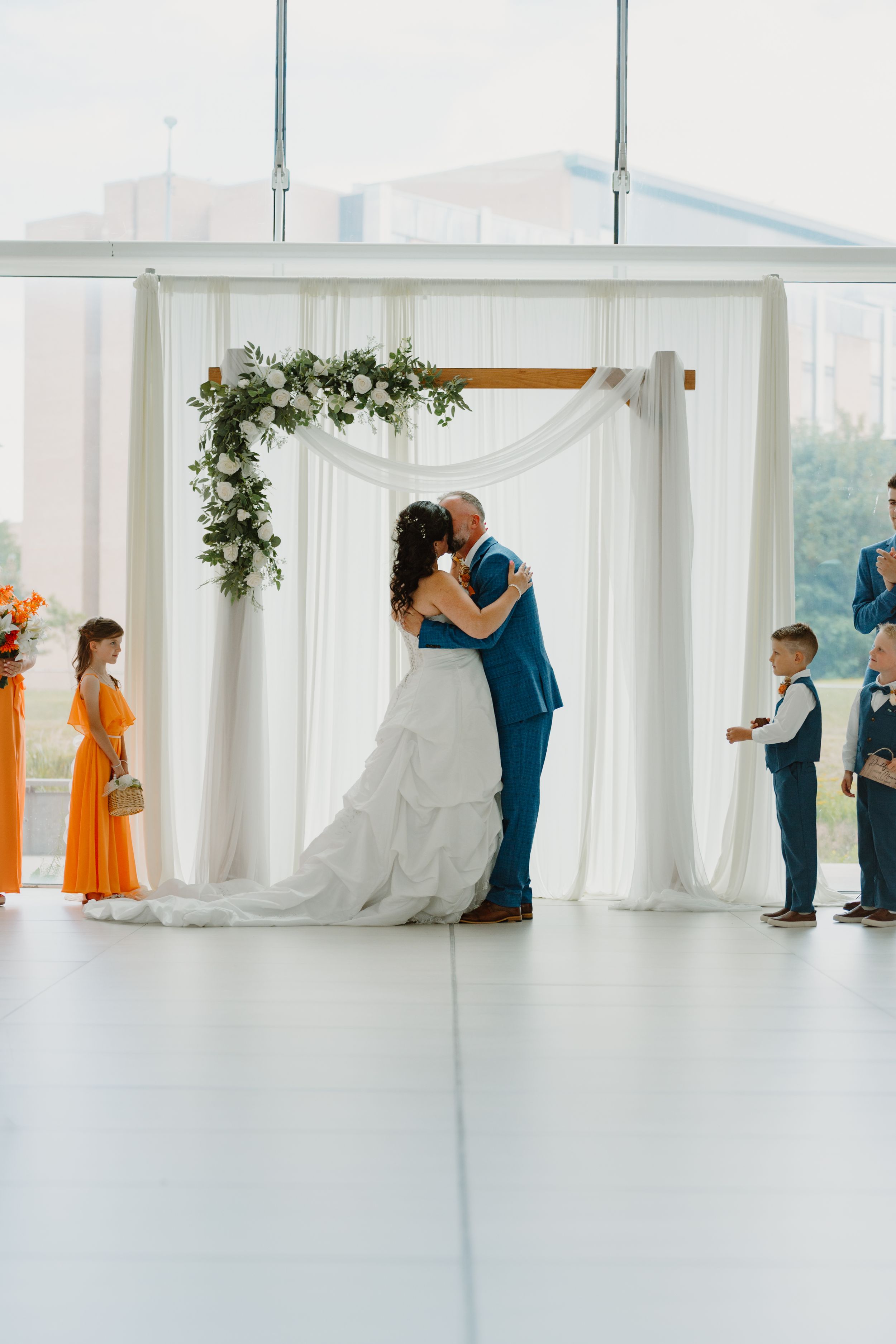 Bride and groom share first kiss under floral arch with white draping, flanked by wedding party in orange and blue.