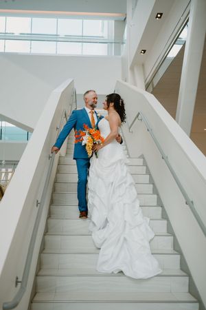 Bride and groom walk down white staircase, she holds orange bouquet, he wears blue suit.