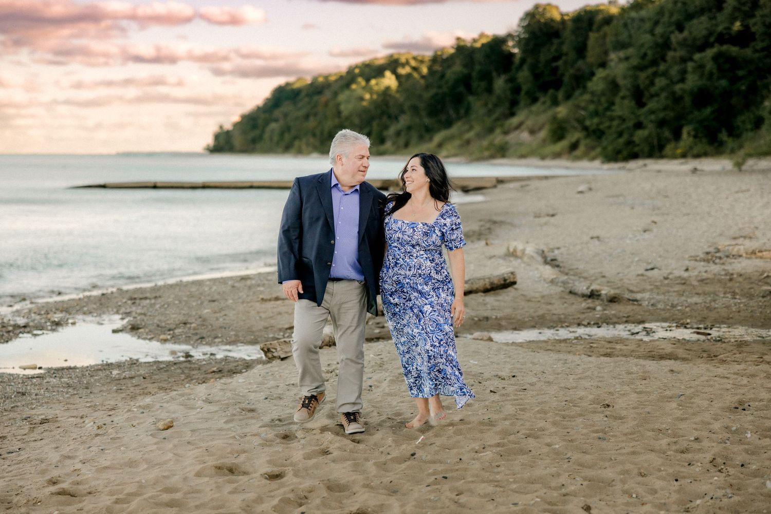 A couple walks together on a sandy beach at sunset with wooded cliffs in the background.