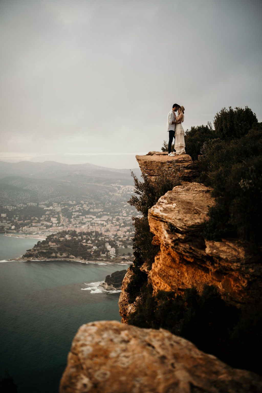 A couple stands embracing on a dramatic clifftop overlooking a coastal town.