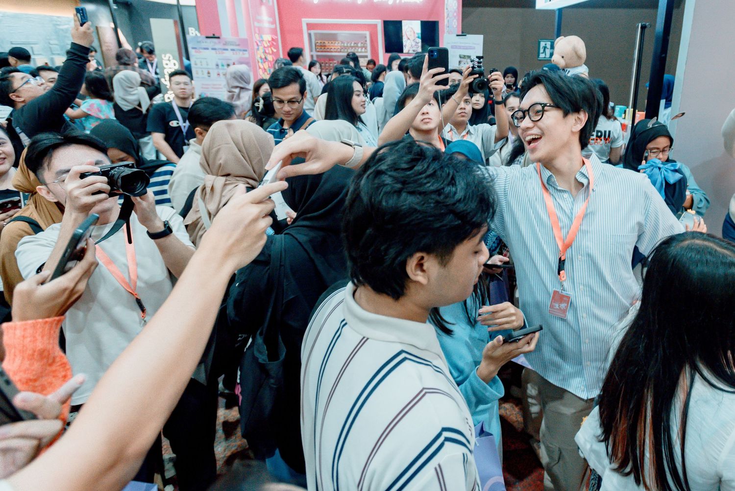 A crowded indoor event with people holding up phones and cameras in an energetic atmosphere with pink lighting.
