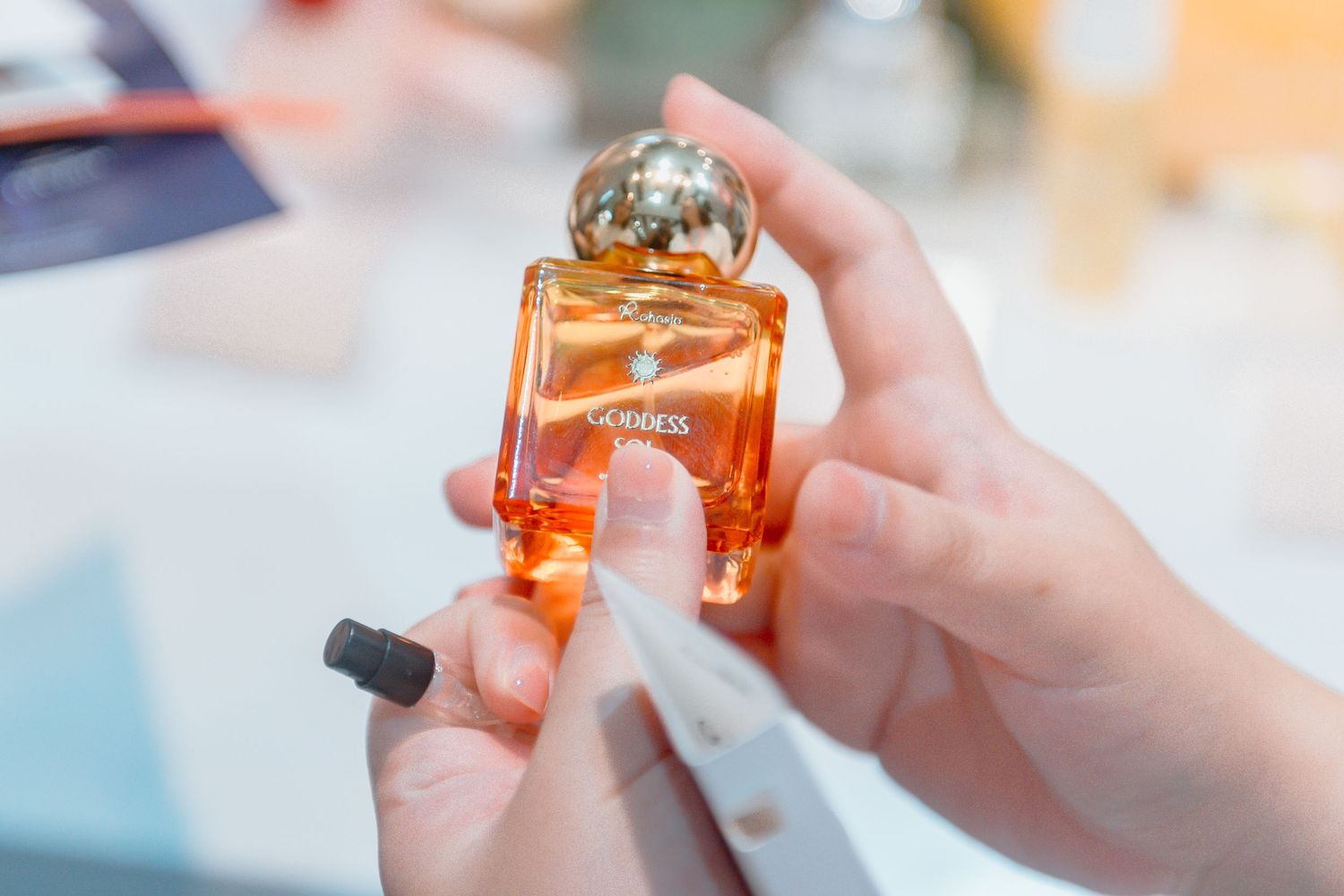 Hands holding and examining a bottle of orange perfume in natural lighting against a blurred background.