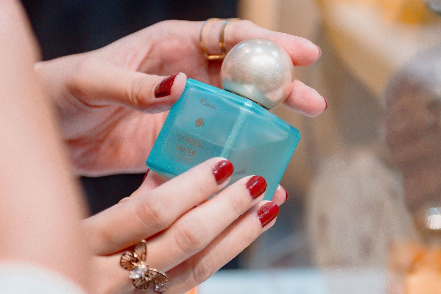 Hands with red manicured nails hold a turquoise perfume bottle with a pearl-like cap against a blurred background.