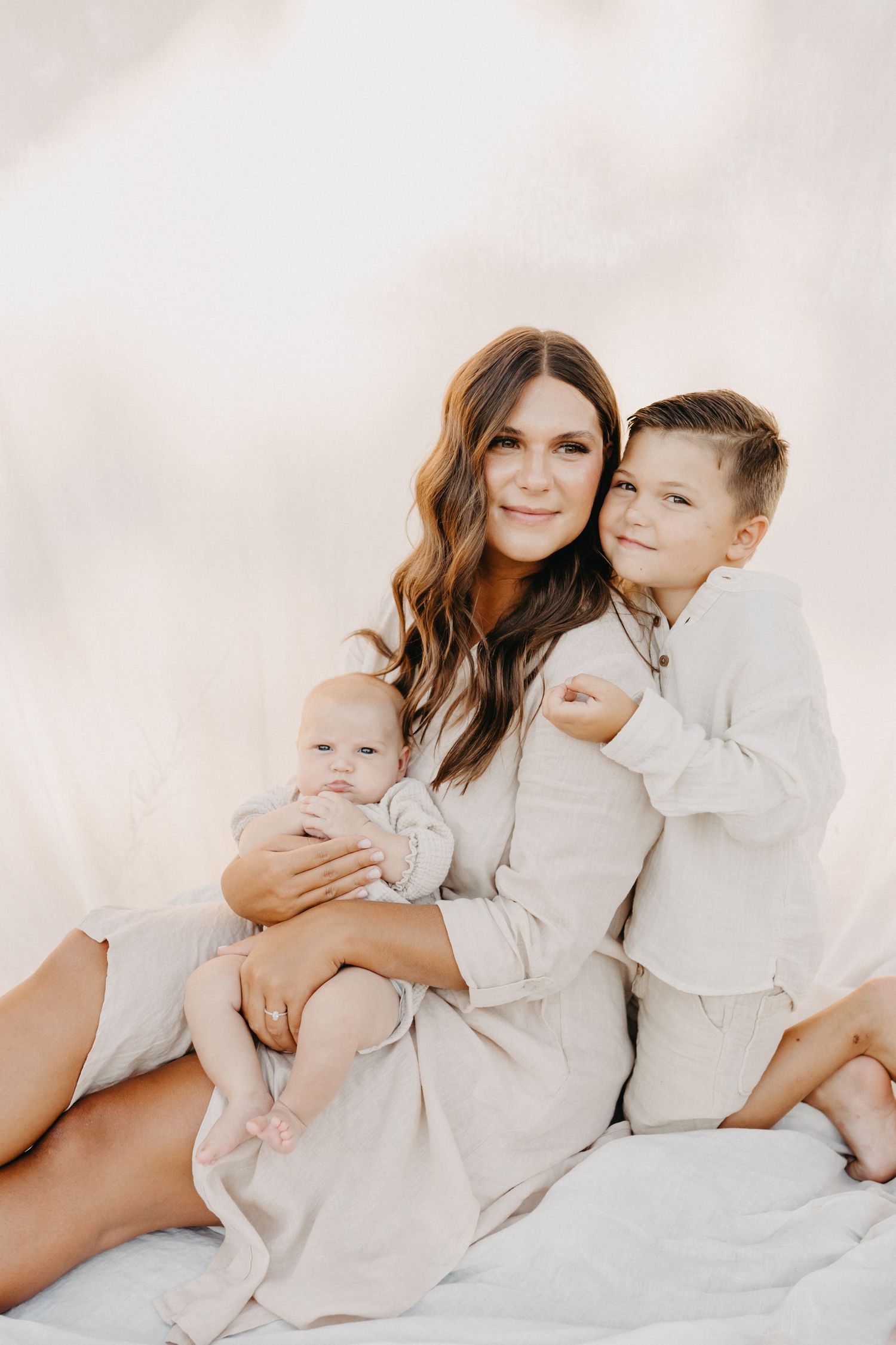 A mother with long brown hair sits holding a baby while an older boy hugs her, all wearing neutral beige tones.