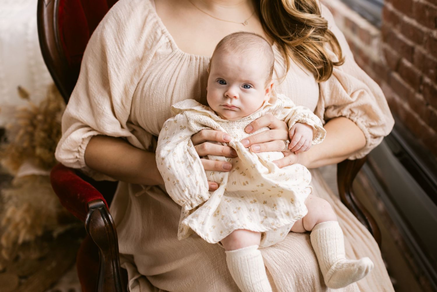 Blue eyed baby girl wearing a cream floral dress is held by her mother wearing a cream feminine dress in an antique chair in North Andover.