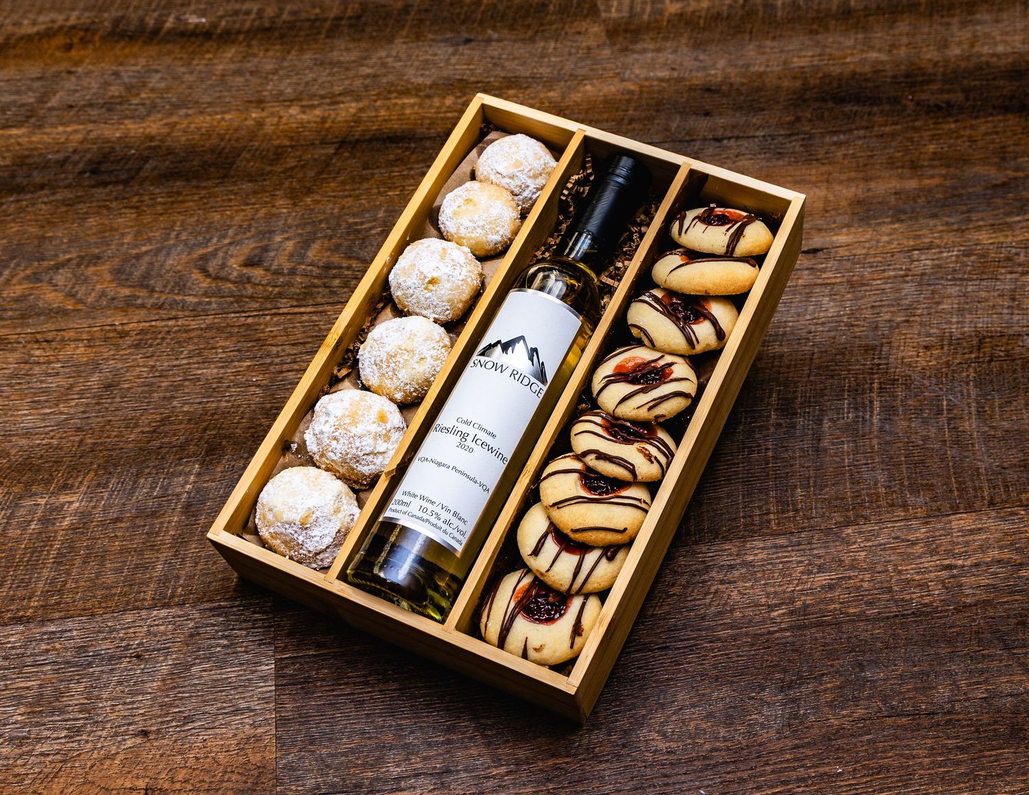 Wooden gift box with compartments displaying artisanal chocolates and wine bottle on rustic wooden surface.