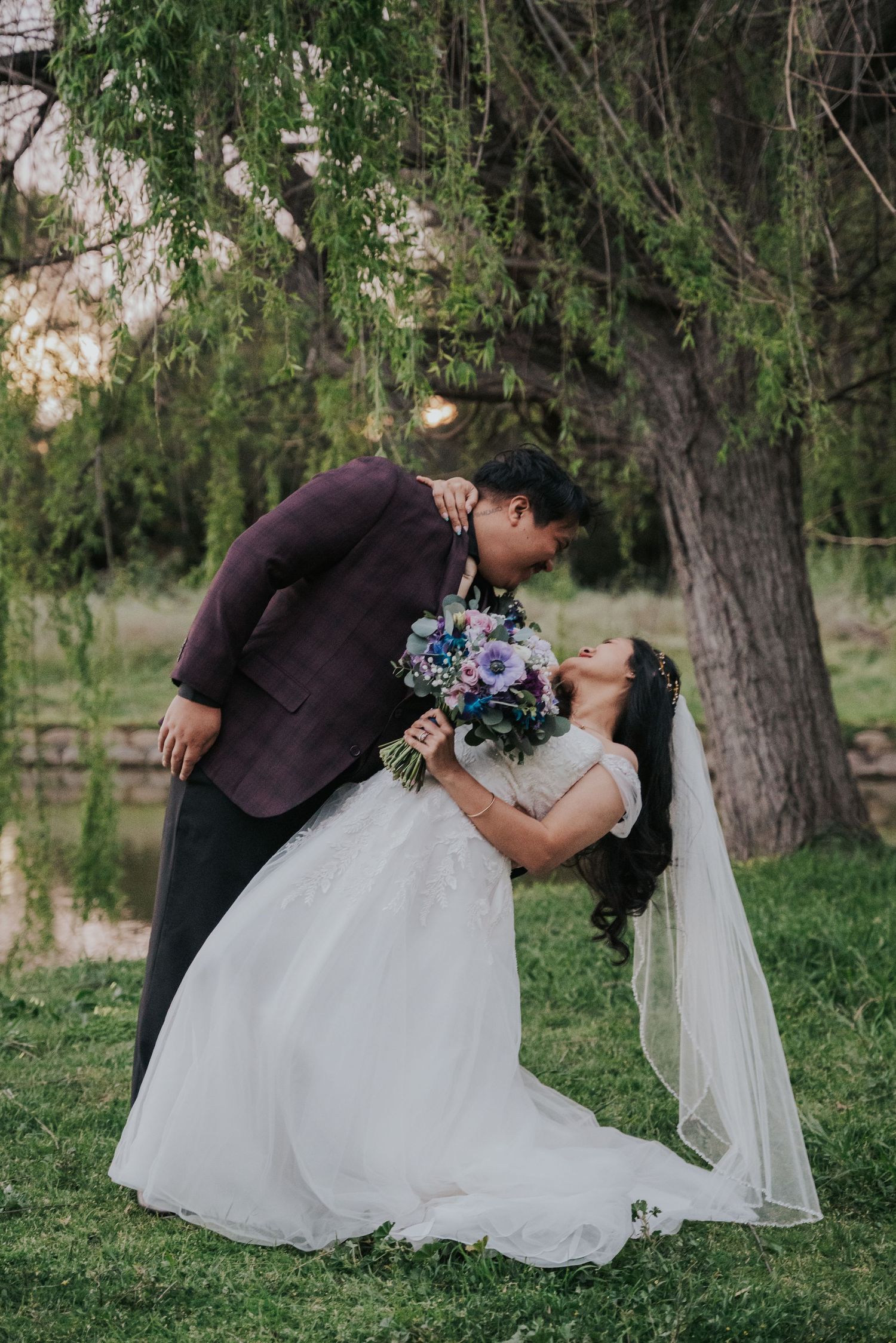 A romantic wedding couple shares a dip kiss pose in a lush green garden setting.