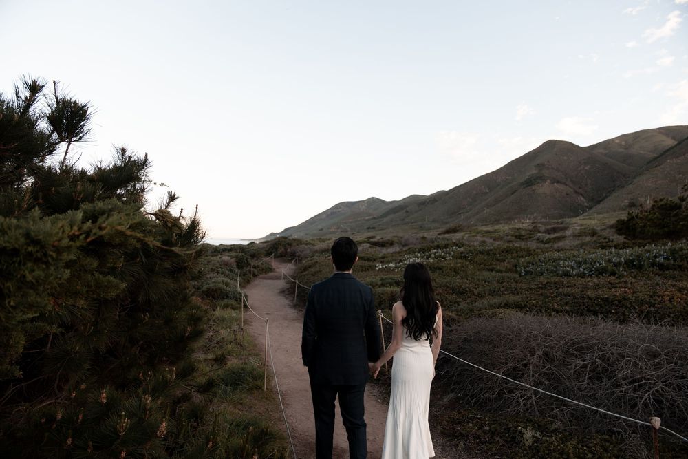 Couple in wedding attire walking along scenic mountain path during sunset at Golden Gate Bridge overlook.
