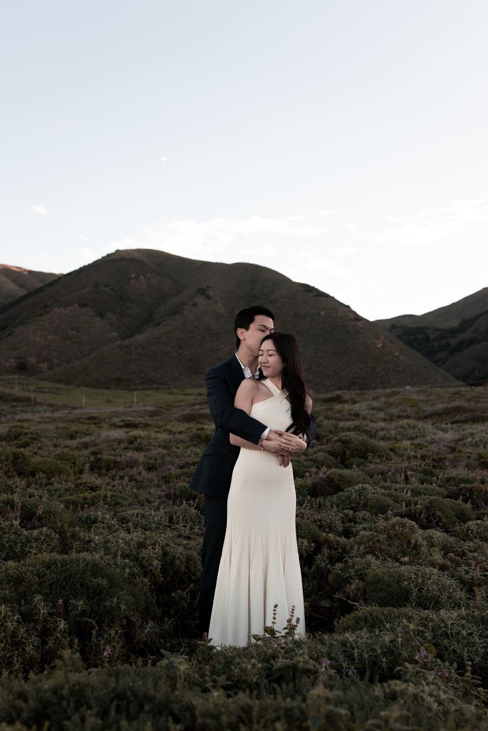 A romantic wedding couple embraces against a backdrop of rolling hills and mountains at dusk.