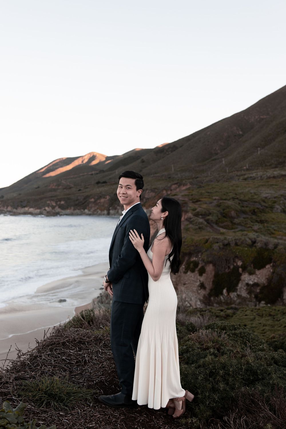 A couple shares a romantic moment against a dramatic coastal landscape with mountains and ocean at sunset.