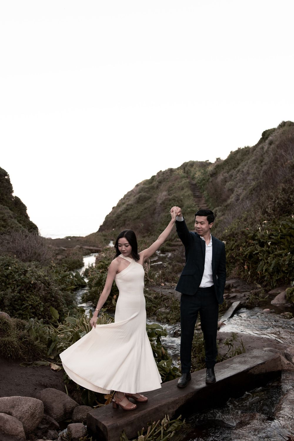 A romantic couple in wedding attire poses together on rocky coastal terrain at sunset.