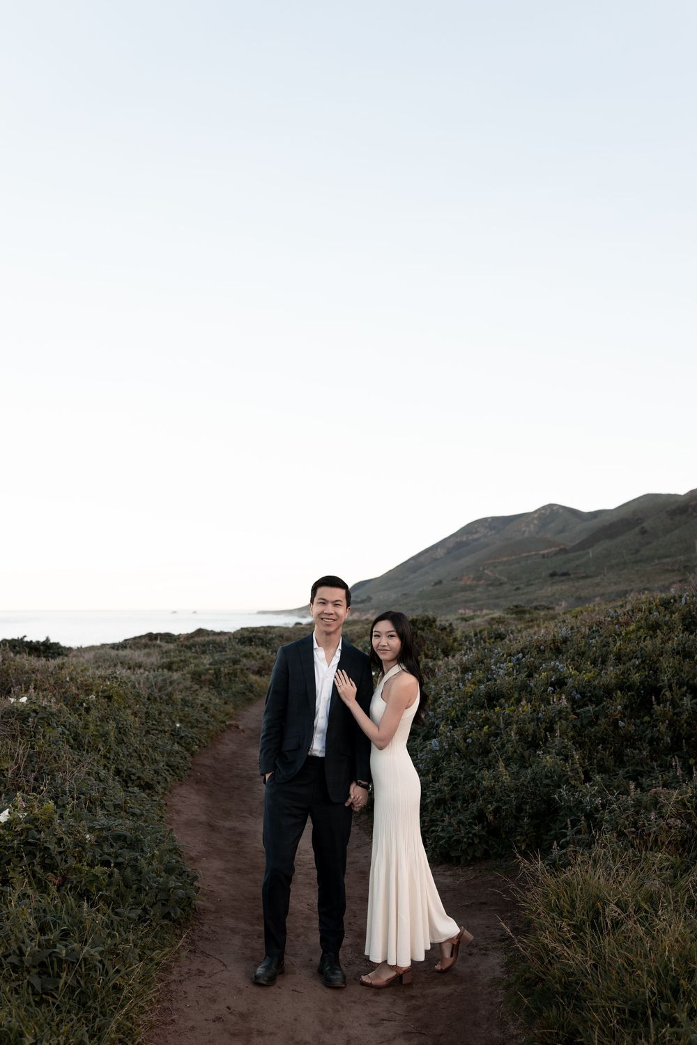 A couple shares a romantic moment on a scenic mountain trail at dusk against a dramatic natural landscape.