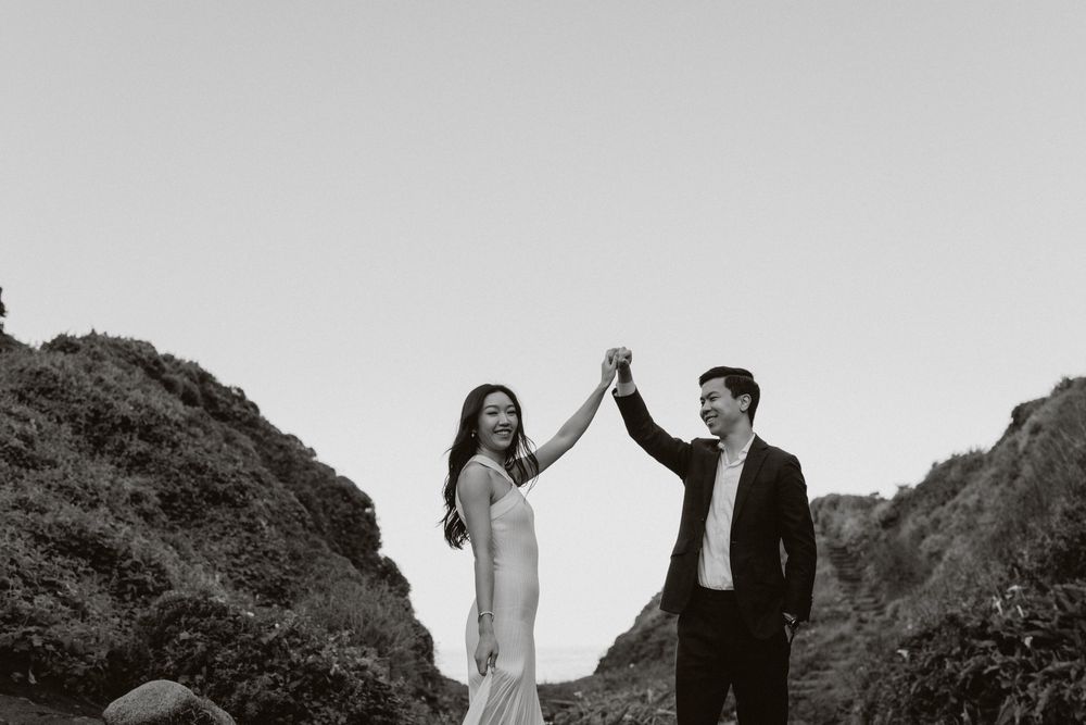 Couple in formal wedding attire walking together on a scenic mountain path at sunset.