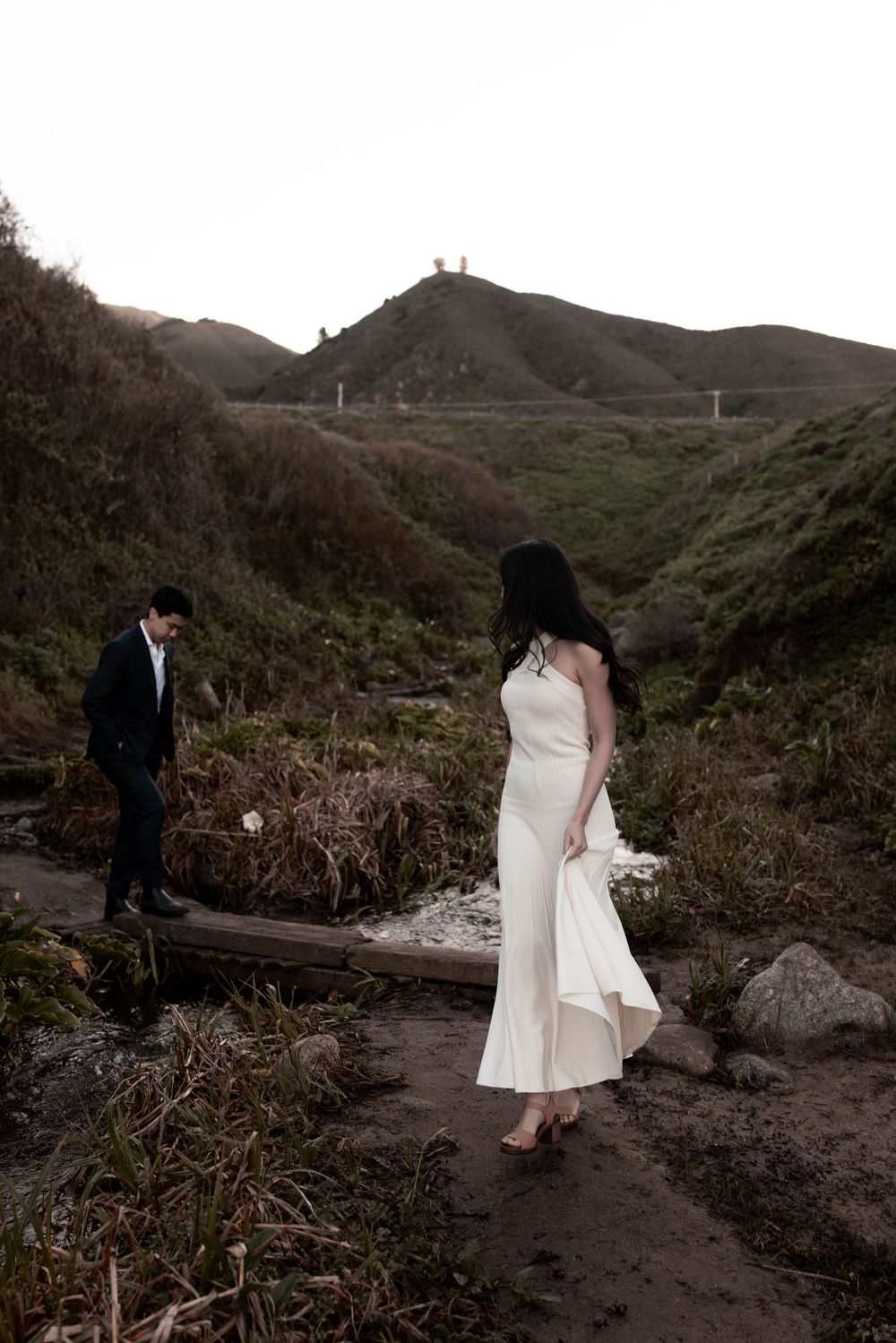 A couple in formal wedding attire walking along a dirt path with rolling hills and mountains in the background at dusk.