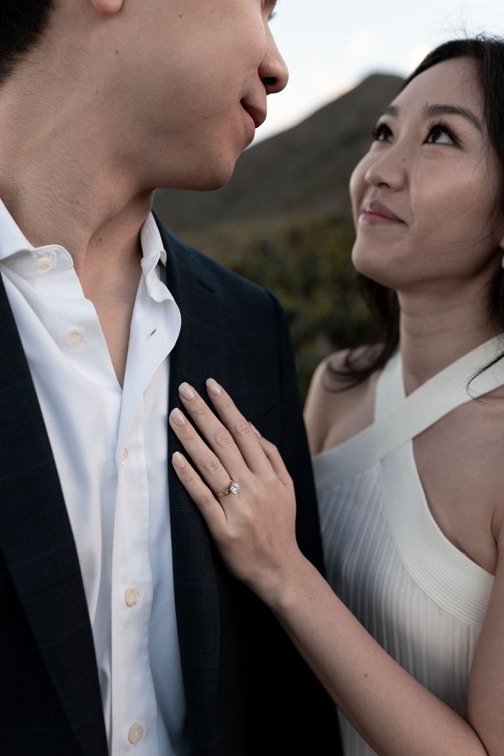 A romantic engagement photo showing a couple in formal attire gazing at each other against a mountain backdrop.