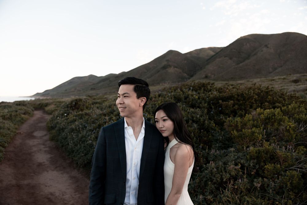 A couple stands together on a scenic mountain trail with green brush and rolling hills in the background.