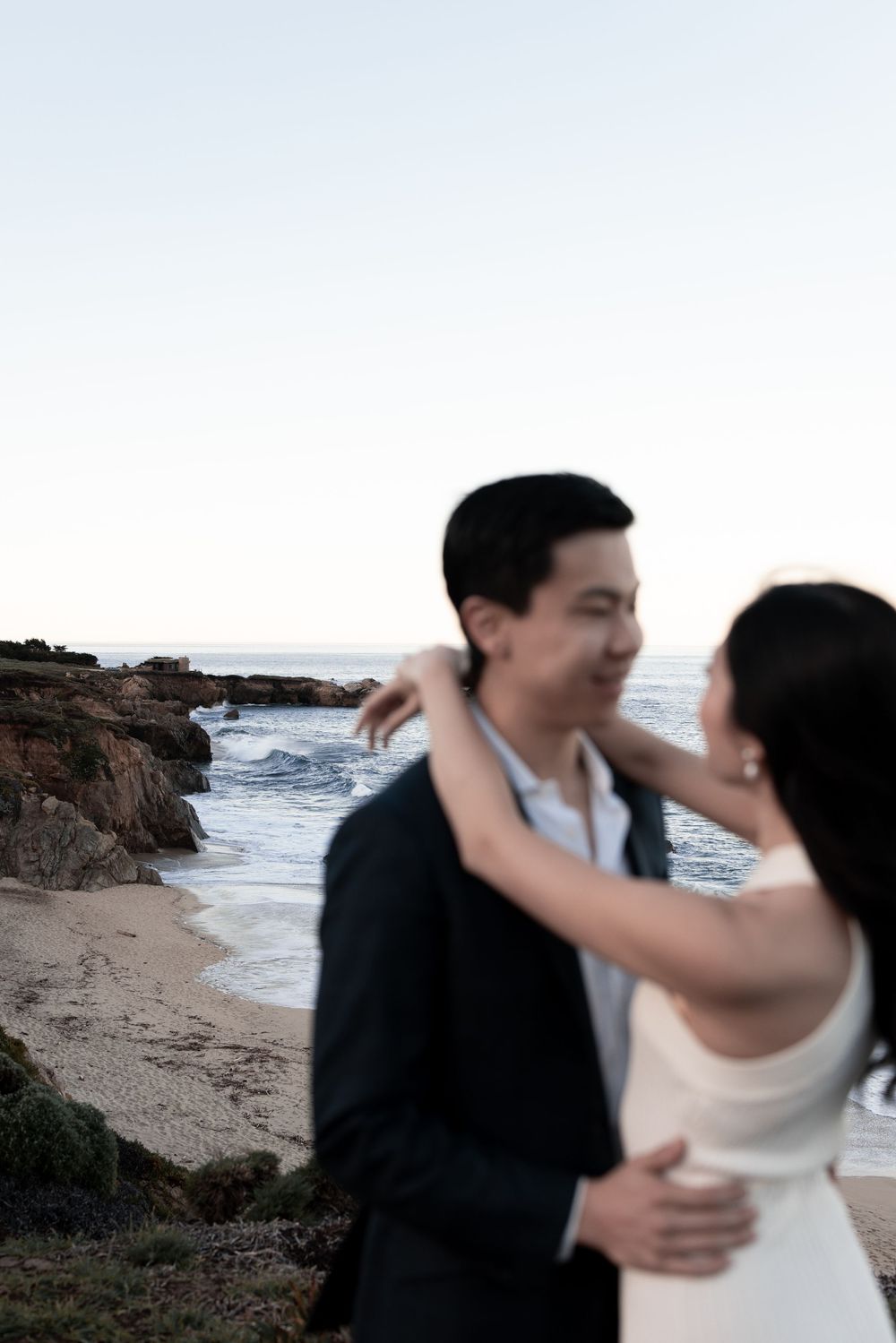 A couple in wedding attire dances and embraces on a rocky beach with ocean waves in the background.