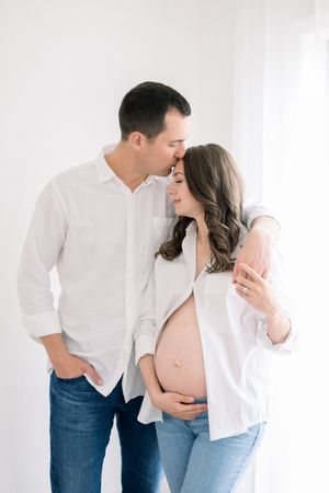 A tender maternity photo of a couple in white shirts and jeans embracing against a light background at a studio session in northern virginia
