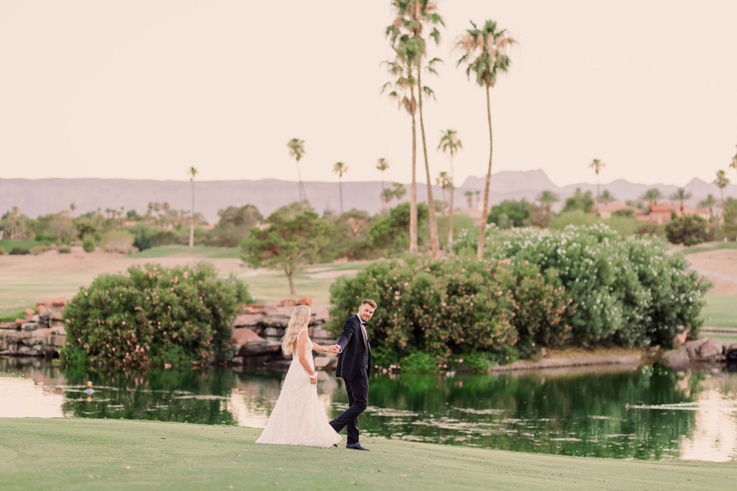 A couple in formal wedding attire walks together through a lush garden path surrounded by trees at Canyon Gate Country Club in Las Vegas.