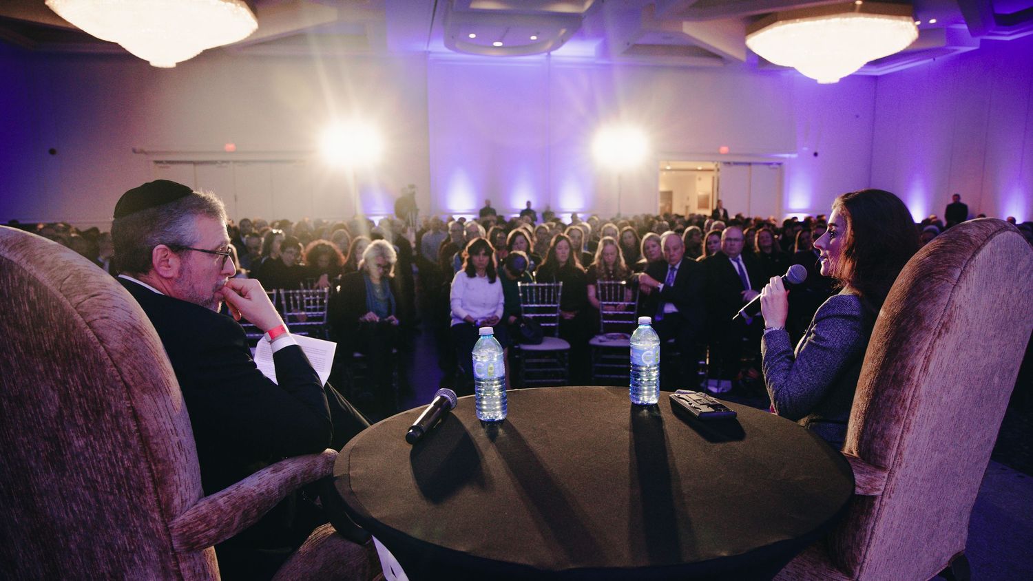 Crowded event venue with purple lighting and people gathered around tables during a conference or meeting.