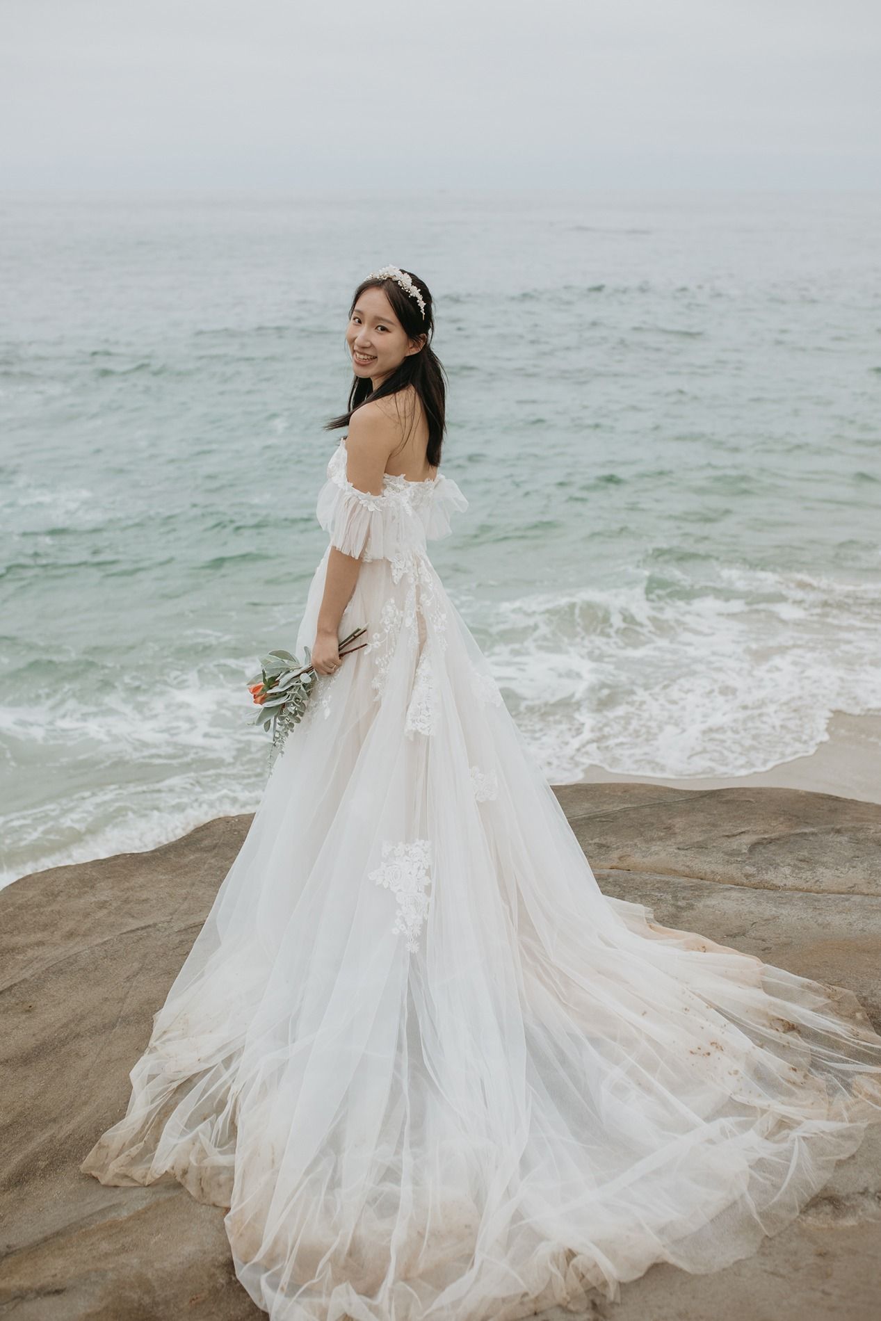 A flowing white wedding dress with a dramatic train billows in the ocean breeze on a rocky beach shoreline at Windansea Beach in La Jolla.