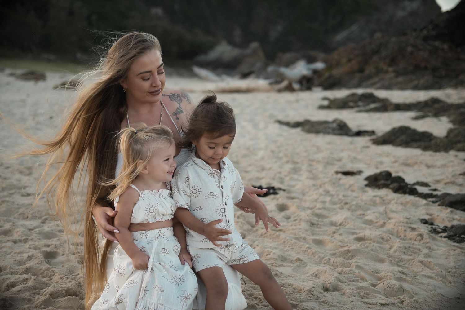 A mother with long hair sits on a sandy beach with her two young children in white outfits.