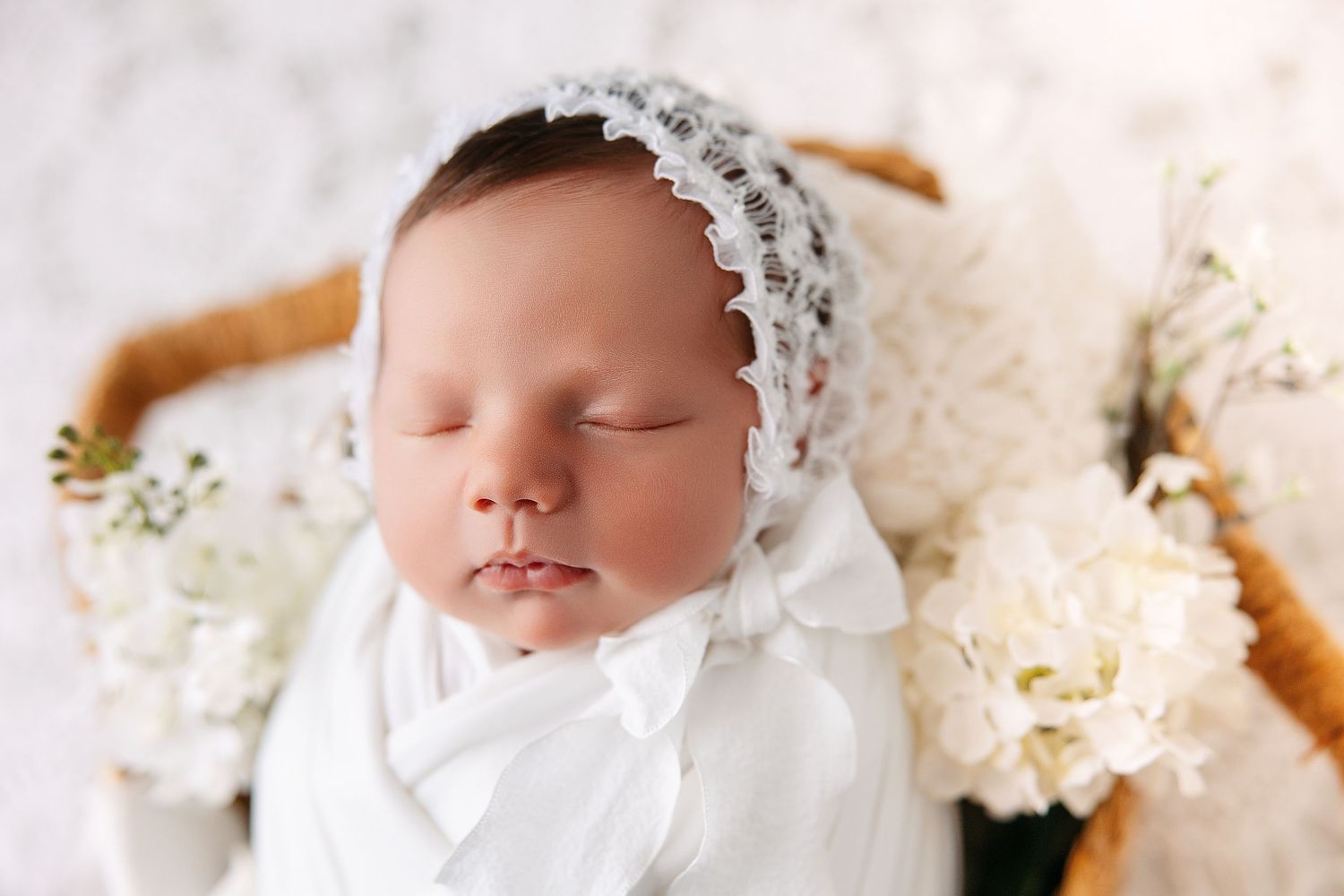 A sleeping baby wearing a white lace bonnet lies on a white blanket surrounded by white flowers.